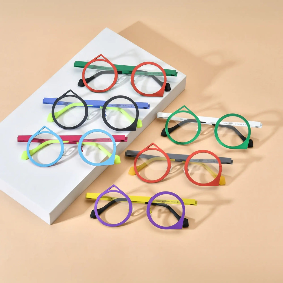 Top-down overview of all six color variants of VoyueLens VL185801 geometric titanium eyeglasses displayed on a beige background, showcasing red-green, purple-yellow, red-grey, green-white, blue-pink, and black-blue artistic polygonal frames.